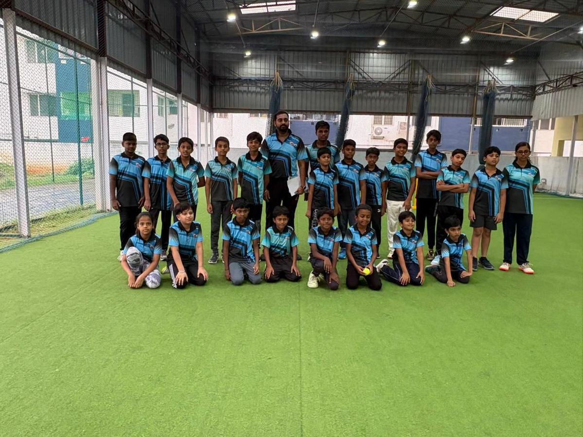 Junior players in teal and black Elite kit standing and kneeling on the indoor turf at the Chennai academy, with practice nets behind them.